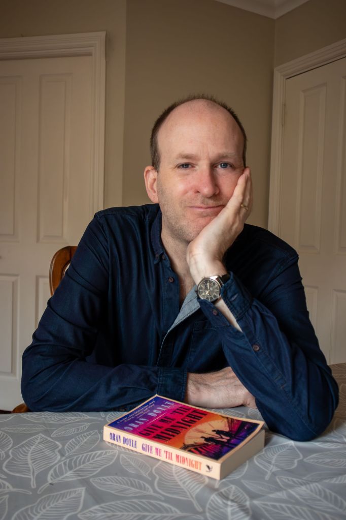 Author sits at table looking at camera with his book in front of him
